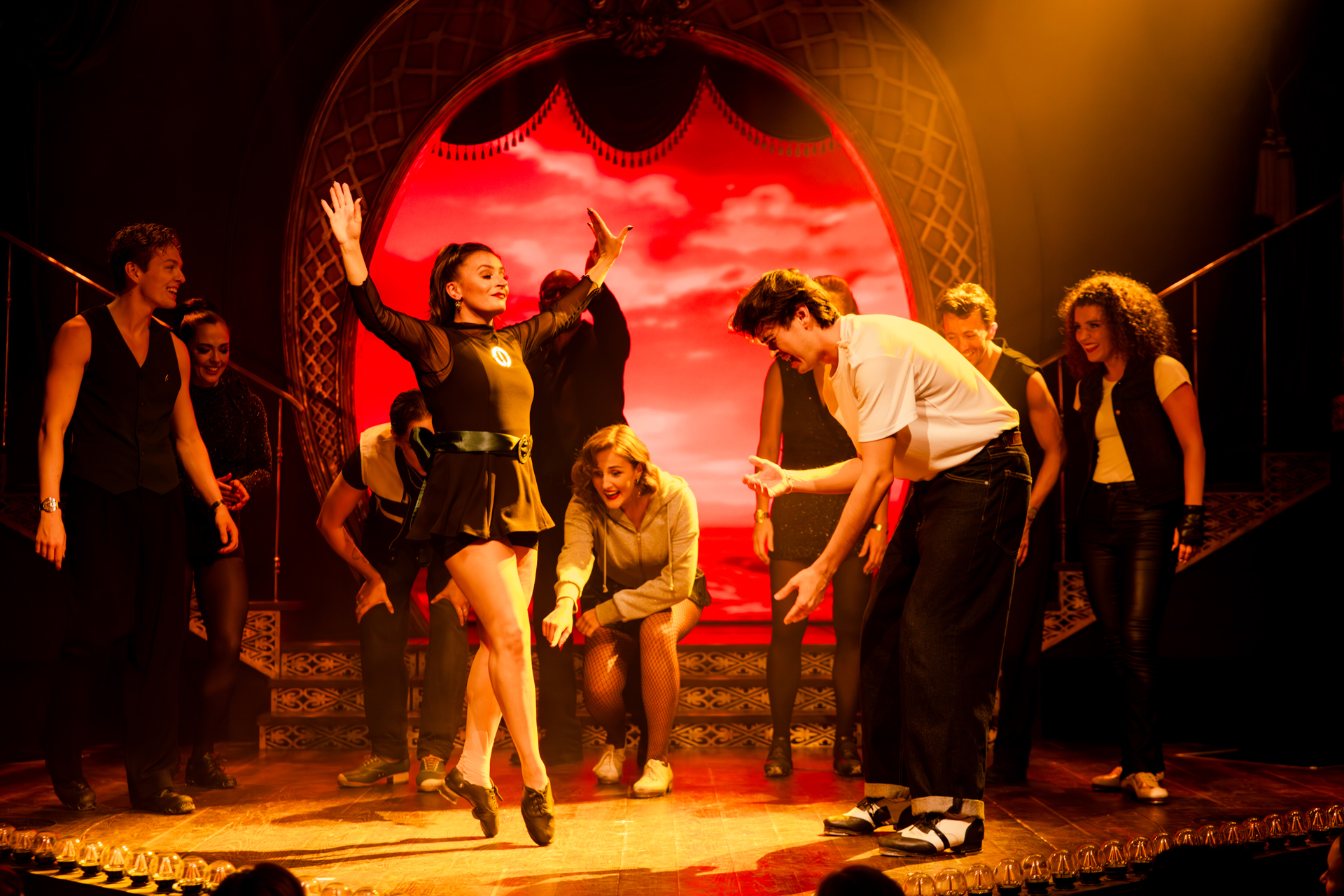 An Irish dancer performers to around 10 others as part of Adam Garcia's Emerald Storm in front of a red background