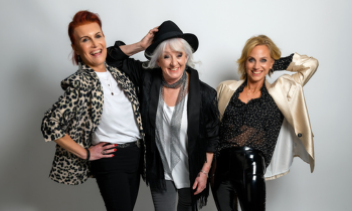 Promotional photo of the musical comedy trio Fascinating Aïda coming to Halifax Victoria Theatre in 2027. Dressed in elegant outfits, the three performers are posed together in a theatrical setting with warm, dramatic lighting.